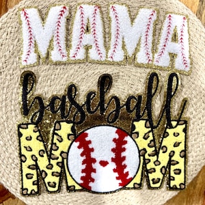 Pode incluir: Um letreiro de mãe de beisebol com glitter dourado com uma bola de beisebol branca e vermelha, uma bola de beisebol dourada com glitter e um letreiro "MAMA" branco e vermelho com glitter dourado.