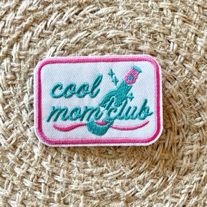 May include: Embroidered patch with the words "cool mom club" in teal script and a pink border. The patch features a teal cocktail with pink accents and sparkles. The patch is rectangular and sits on a woven surface.