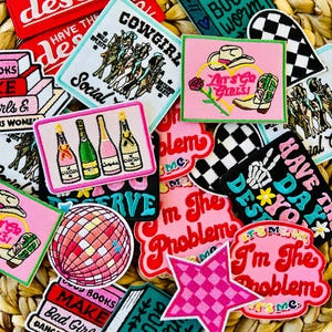 Könnte beinhalten: Eine Sammlung bunter Bügelbilder mit verschiedenen Designs, darunter ein Bücherwurm, ein Cowboy, eine Discokugel und ein Stern. Die Patches haben Sätze wie "Have the day you deserve", "Let's go girls", "I'm the problem" und "Book worm".