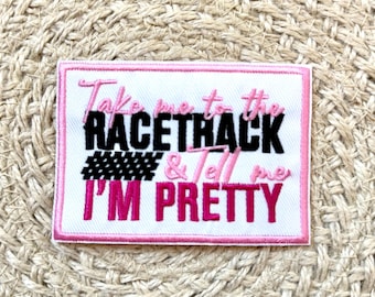 Puede incluir: Parche rectangular con fondo blanco y borde rosa. El texto "Take me to the racetrack & Tell me I'm Pretty" está bordado en negro y rosa. También hay un diseño a cuadros en blanco y negro.