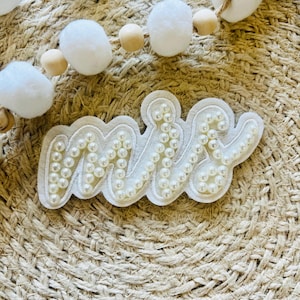 Pode incluir: Remendo de tecido branco com a palavra "Mrs." escrita em contas de pérola. O remendo está sobre um fundo marrom tecido.