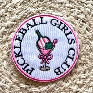 May include: An embroidered patch in pink and white with the text "Pickleball Girls Club" in black lettering. The patch features a pink and white striped cocktail glass with a green lime wedge and a pink bow.