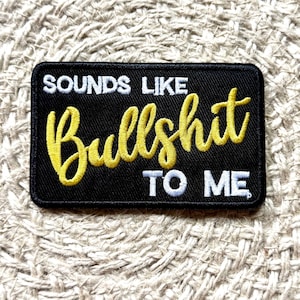 Puede incluir: Un parche rectangular negro bordado con hilo blanco y amarillo. El texto dice SOUNDS LIKE en blanco, Bullshit en una letra cursiva amarilla grande y TO ME en blanco. El parche tiene un borde negro cosido y un acabado de tela texturizada.