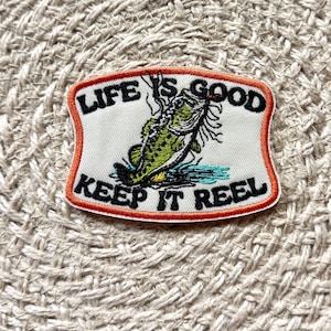 Puede incluir: Un parche bordado con fondo blanco hueso y borde naranja. Presenta un gráfico de un pez verde, agua azul y el texto en negro que dice LIFE IS GOOD KEEP IT REEL. El parche tiene los bordes superior e inferior curvados.