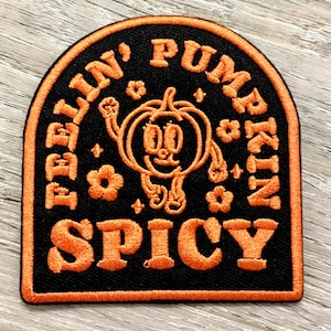 May include: Black and orange embroidered patch with the words "BELLIN' PUMPKIN SPICY" in a curved design. A cartoon pumpkin character is in the center, surrounded by flowers and stars. The patch has an orange border.