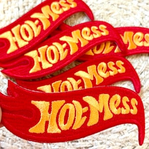 Pode incluir: Vários patches bordados vermelhos com o texto "Hot Mess" em amarelo. Os patches têm um design em forma de chama. Adequados para roupas ou acessórios, adicionam um toque ousado.