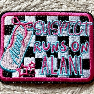 May include: Embroidered patch with a pink border and a black and white checkered background. The patch features a pink and blue sneaker design and the text "SUSPECT RUNS ON ALANI" in pink and blue lettering.