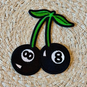 Könnte beinhalten: Gestickter Aufnäher mit zwei schwarzen Kirschen, jeweils mit einer weißen Acht und grünen Stielen und Blättern. Der Aufnäher hat einen schwarzen Rand und ist zum Aufbügeln oder Aufnähen auf Stoff vorgesehen.