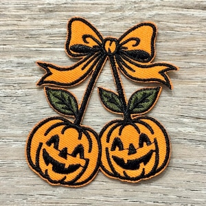 Puede incluir: Parche bordado con dos calabazas naranjas con caras sonrientes, conectadas por tallos y hojas verdes, rematadas con un lazo naranja con ribete negro. El parche está diseñado para Halloween.