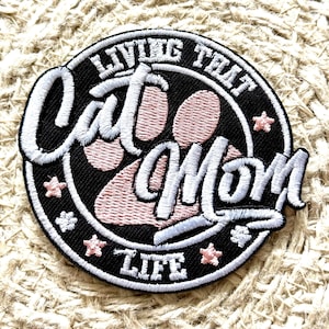 May include: A black and white embroidered patch with a pink paw print in the center. The text around the paw print reads "Living That Cat Mom Life".