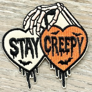 May include: Embroidered patches in the shape of hearts with the words "STAY CREEPY" in black. One heart is cream-colored, the other is orange. A skeleton hand is holding the hearts. Bats and dripping details complete the design.