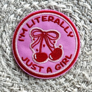 May include: A circular embroidered patch with a pink background and a red border. The center features two red cherries tied with a red bow. The text "I'M LITERALLY JUST A GIRL" is embroidered in red capital letters around the circular edge.