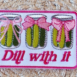 May include: Embroidered patch featuring three jars of pickles with pink bows, set against a white background with a pink border. The phrase "Dill with it" is embroidered in pink script below the jars.