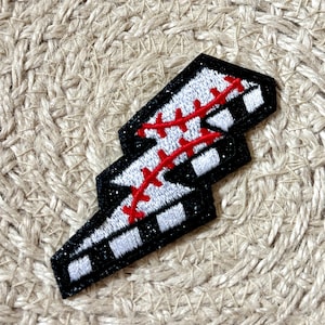 May include: A baseball-themed patch in the shape of a lightning bolt. The patch features a black border with glitter, white and black squares, and red stitching, resembling a baseball. The patch is on a textured, beige surface.