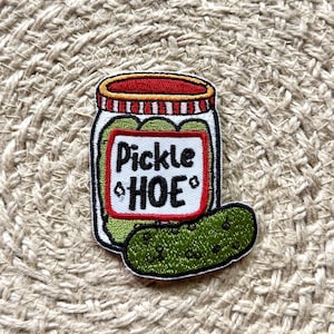 May include: An embroidered patch featuring a glass jar filled with green pickles, topped with a red and white striped lid. A white label on the jar has a red border and the black text "Pickle HOE" with small diamond icons. A single green pickle sits in front.