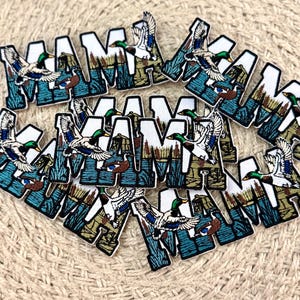 May include: A collection of embroidered patches featuring the word MAMA in bold white block letters. Each letter contains a detailed outdoor scene with flying mallard ducks, blue water, and brown cattails against a textured background.