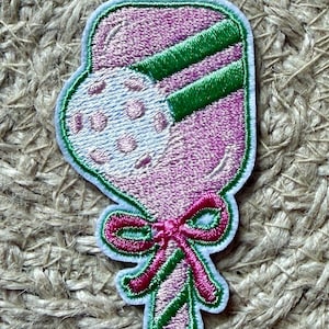 May include: Embroidered patch in the shape of a pink and white lollipop with green stripes and a pink bow. The patch has a white border and is likely for clothing or accessories.