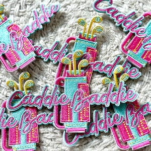 Op de afbeelding: Geborduurde patches met een golfthema. Elke patch heeft een golftas in turquoise en roze, met de tekst "Caddie Baddie" in een cursief lettertype. De golfclubs zijn geel en groen. De patches liggen op een getextureerd, lichtgrijs oppervlak.