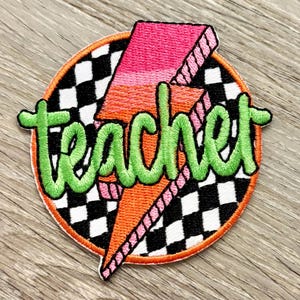 Puede incluir: Parche bordado con la palabra "teacher" en escritura verde. El parche presenta un diseño de rayo rosa y naranja sobre un fondo a cuadros blanco y negro, todo dentro de un borde circular naranja.