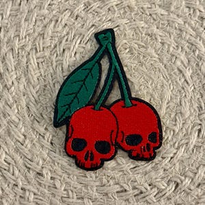 May include: A fabric patch featuring two red cherry-shaped skulls with black eye sockets and nose cavities. The cherries are connected by green stems and a single green leaf with black stitched detailing, all outlined in a thick black border.