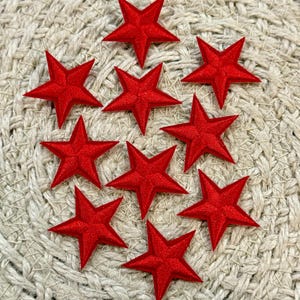 May include: Nine red star-shaped fabric appliques arranged on a woven beige surface. Each star has five points and a slightly raised, textured appearance. The stars are evenly spaced and create a visually appealing pattern.