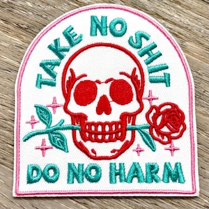 Może przedstawiać: Haftowana naszywka z białym tłem, różową obwódką oraz turkusowymi i czerwonymi akcentami. Naszywka przedstawia czerwoną czaszkę z różą w ustach oraz napisy "TAKE NO SHIT" i "DO NO HARM" w turkusowych literach.