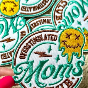 Op de afbeelding: Geborduurde patches met de woorden "Moms Club" en "Overstimulated" in een cirkelvormig ontwerp. De patches hebben teal en bruine stiksels op een witte achtergrond, met een gele, smeltende gezichtsafbeelding. De patches zijn waarschijnlijk voor kleding of accessoires.