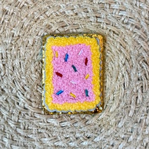May include: An embroidered patch of a pink and yellow pop tart with colourful sprinkles. The patch is on a woven background.