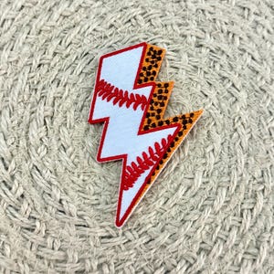Könnte beinhalten: Ein gestickter Aufnäher in Form eines Blitzes. Die linke Seite zeigt ein weißes Baseball-Muster mit roten Nähten, während die rechte Seite ein braun-schwarzes Leopardenmuster aufweist. Der gesamte Blitz ist mit rotem Garn umrandet.