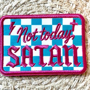 May include: Embroidered patch with a magenta border and the phrase "Not today SATAN" in magenta gothic lettering. The background is a checkered pattern of white and turquoise squares, with a lightning bolt and cross symbol.