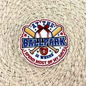 May include: A circular embroidered patch featuring a white baseball with a red bow, crossed wooden bats, and a brown baseball glove. The text reads AT THE BALLPARK IS WHERE I SPEND MOST OF MY DAYS in blue and red lettering on a white background.