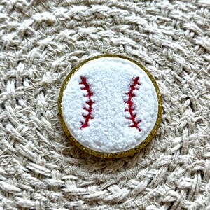 Könnte beinhalten: Ein weißer Baseball mit roten Nähten, umgeben von einem goldenen Glitzerrand. Der Baseball liegt auf einem gewebten Hintergrund.