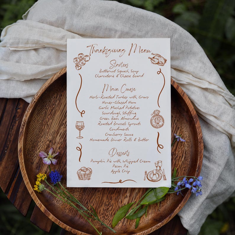 May include: A Thanksgiving menu printed on a cream-colored card, featuring a brown decorative border and text. The menu lists starters, main courses, and desserts, including butternut squash soup, turkey, and pumpkin pie. The card is placed on a wooden tray with floral accents.