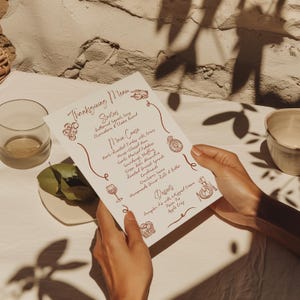 May include: A Thanksgiving menu printed on cream-colored paper, held by hands. The menu features a handwritten style with decorative illustrations and lists starters, main courses, and desserts. The menu includes dishes like herb-roasted turkey and pumpkin pie.
