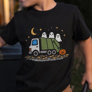 May include: A black short-sleeve t-shirt featuring a graphic of a green garbage truck with three white ghosts riding in the back. The design includes a yellow crescent moon, small stars, and an orange jack-o'-lantern on the ground.