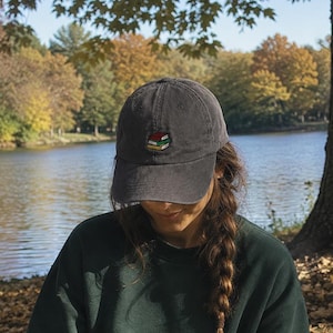 May include: A charcoal grey baseball cap featuring a small embroidered stack of books on the front panel. The cap has a curved brim and a structured crown, constructed from a cotton-like fabric with visible stitching details along the panels.