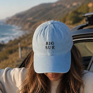 May include: A light blue baseball cap with a curved brim and the text BIG SUR embroidered in black thread on the front panel. The cap features a structured crown with a button on top and stitched eyelets for ventilation.