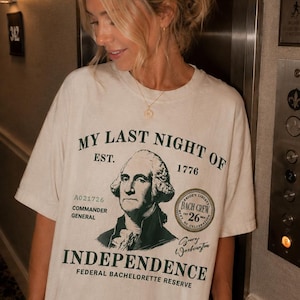 May include: An off-white short-sleeve t-shirt featuring a dark green graphic print of George Washington. The text reads: MY LAST NIGHT OF INDEPENDENCE, EST. 1776, A021726 COMMANDER GENERAL, BACH CREW 26, and FEDERAL BACHELORETTE RESERVE.