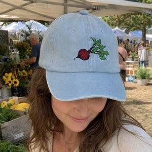 May include: A light blue baseball cap featuring an embroidered graphic of a red beet with green leaves centered on the front panel. The cap has a structured crown, a curved brim, and a button on top, constructed from a denim-like fabric.