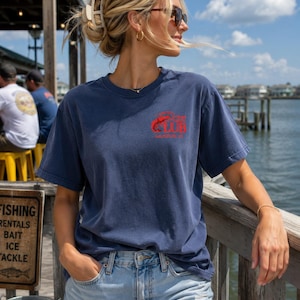 Puede incluir: Una camiseta azul marino de manga corta con un gráfico rojo en el pecho izquierdo que representa un crustáceo y el texto "THE CRAB LUB GALVESTON, TX". La camiseta tiene cuello redondo y un corte holgado en tejido de algodón liso.