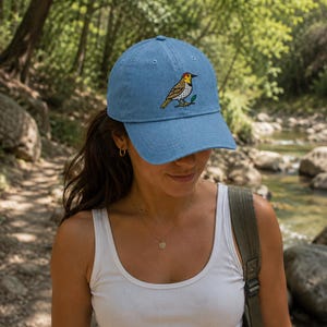 Könnte beinhalten: Eine hellblaue Baseballkappe mit einer gestickten Grafik eines Goldflikers, der einen gelb-braunen Körper, einen roten Fleck am Kopf und einen schwarzen Schnabel hat und auf einem kleinen grünen Zweig in der Mitte der Vorderseite sitzt.