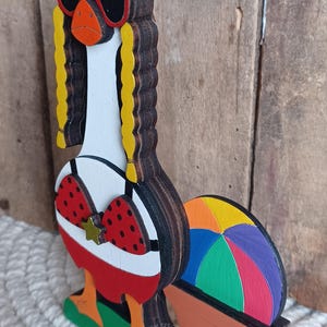 May include: A wooden figurine of a goose wearing sunglasses, a red bikini top with white polka dots, and a gold star. The goose is standing on a green base with a rainbow beach ball behind it. The figurine is made of layered wood and painted in bright colors.