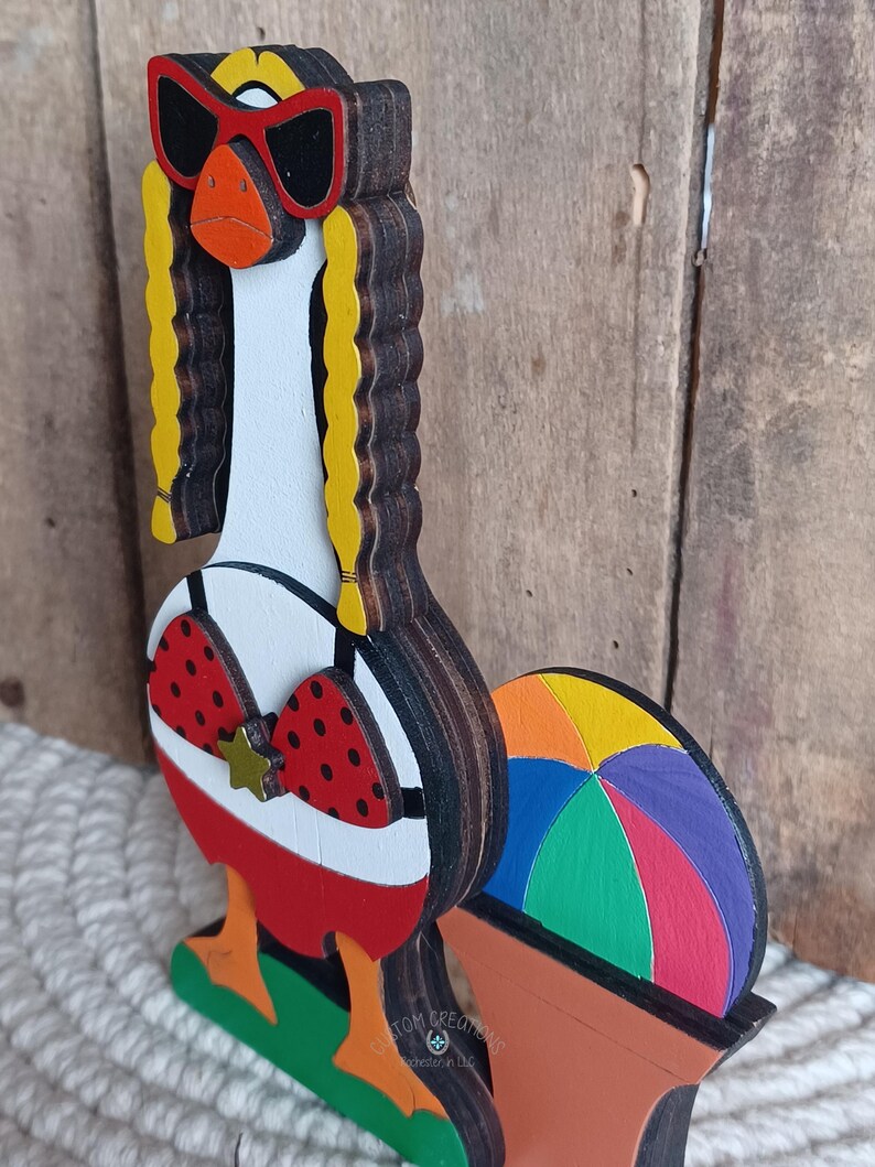 May include: A wooden figurine of a goose wearing sunglasses, a red bikini top with white polka dots, and a gold star. The goose is standing on a green base with a rainbow beach ball behind it. The figurine is made of layered wood and painted in bright colors.
