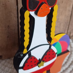 May include: A wooden goose figurine wearing red sunglasses, a red and white polka dot bikini, and holding a beach ball. The goose has yellow hair in pigtails.