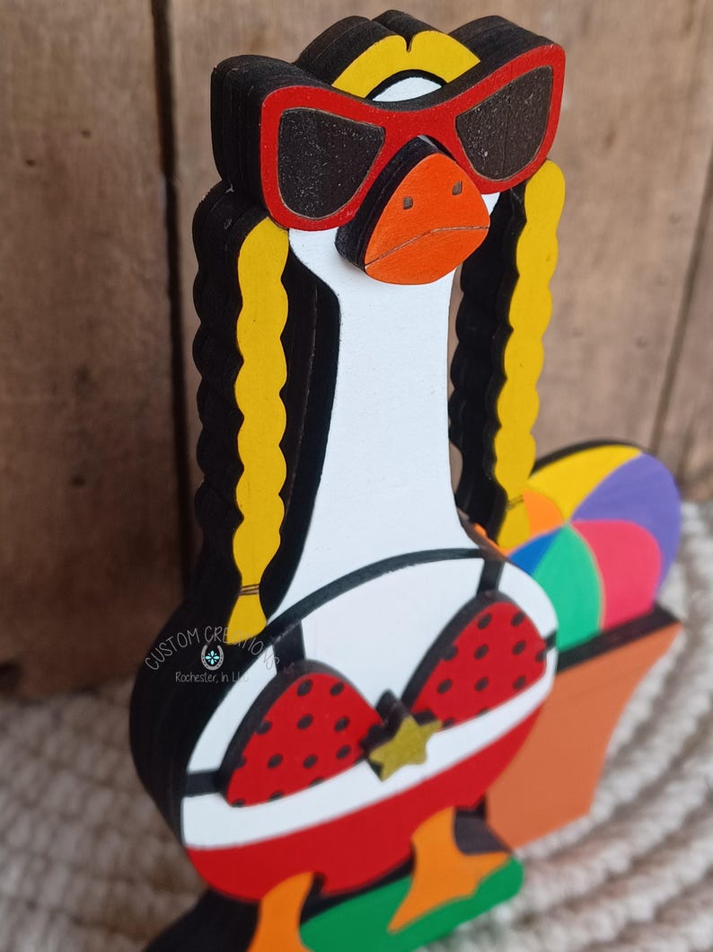 May include: A wooden goose figurine wearing red sunglasses, a red and white polka dot bikini, and holding a beach ball. The goose has yellow hair in pigtails.