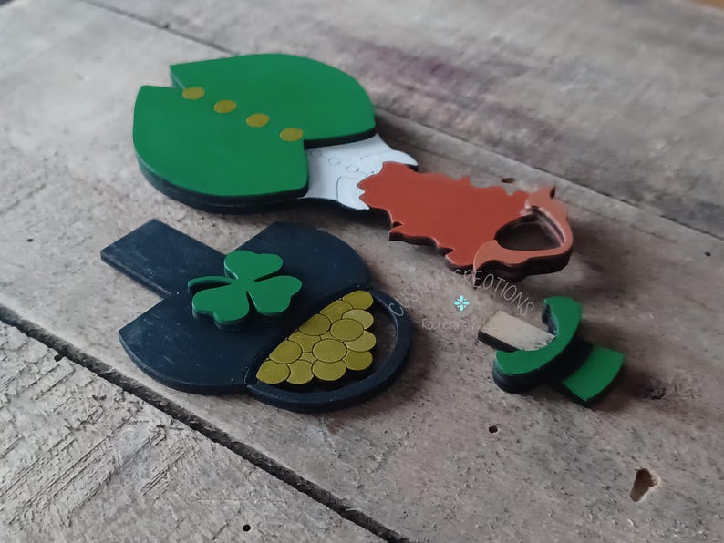 May include: A set of wooden cutouts for a leprechaun, pot of gold, and shamrock. The leprechaun is wearing a green hat and coat, and the pot of gold is filled with gold coins. The shamrock is green.