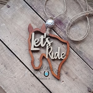 May include: Wooden horse head cutout with the words "Let's Ride" and the text "Custom Creations Rochester, In LLC" below. The horse head is brown and the text is white. A brown and white patterned bead is attached to a brown twine rope.