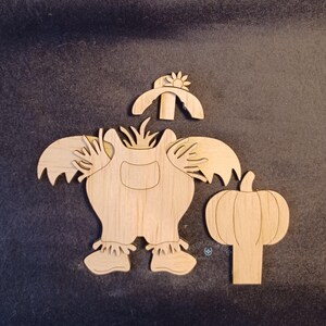 May include: Wooden fall-themed cutouts. The set includes a scarecrow figure with a hat and a pumpkin. The scarecrow has overalls and a sunflower on its hat. The pumpkin has vertical lines.