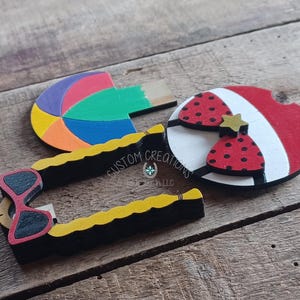 May include: Three wooden cutouts, one shaped like a beach ball with red, yellow, blue, and green colors, one shaped like a pair of sunglasses with black frames and red lenses, and one shaped like a life preserver with red and white stripes and a red bow with black polka dots. The life preserver has a gold star on the bow. The cutouts are painted and have a textured surface. The text "CUSTOM CREATIONS LLC" is visible on the sunglasses cutout.