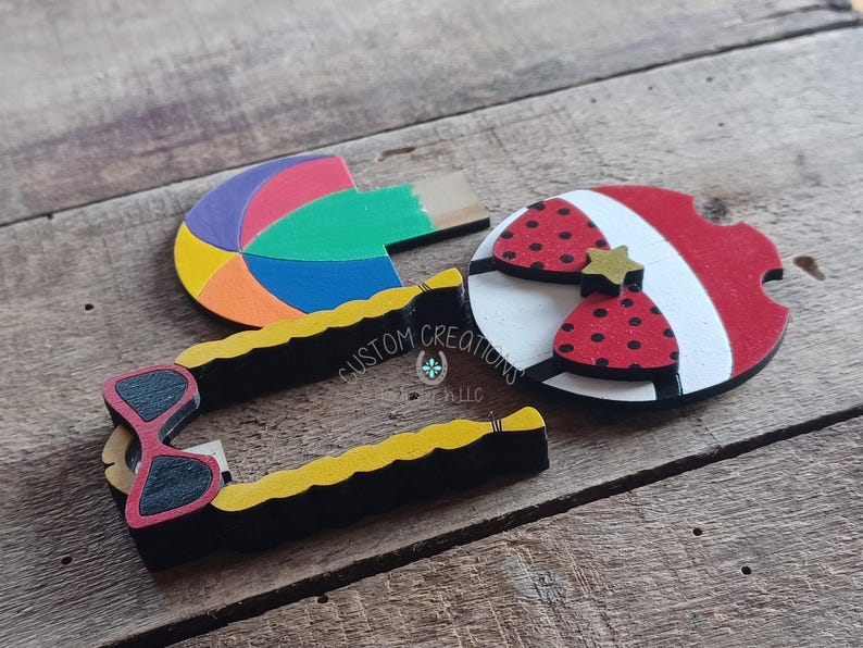 May include: Three wooden cutouts, one shaped like a beach ball with red, yellow, blue, and green colors, one shaped like a pair of sunglasses with black frames and red lenses, and one shaped like a life preserver with red and white stripes and a red bow with black polka dots. The life preserver has a gold star on the bow. The cutouts are painted and have a textured surface. The text "CUSTOM CREATIONS LLC" is visible on the sunglasses cutout.
